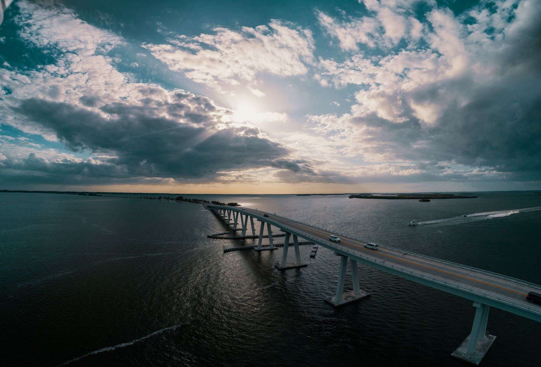 Aerial view of a bridge causeway crossing a wide bay under a cloudy sky, with cars driving across and a boat passing below