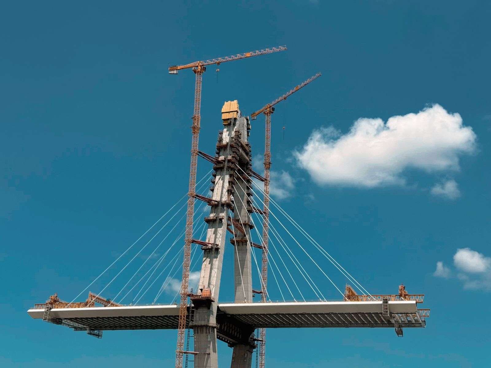 Tall bridge support towers with construction cranes rising against a blue sky, with the bridge deck extending on either side