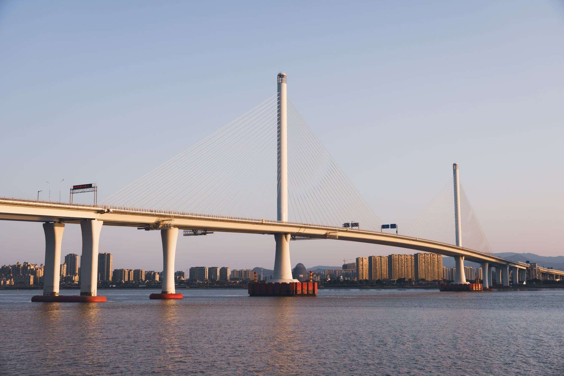A tall cable-stayed bridge under construction spanning a working river with port infrastructure in the background
