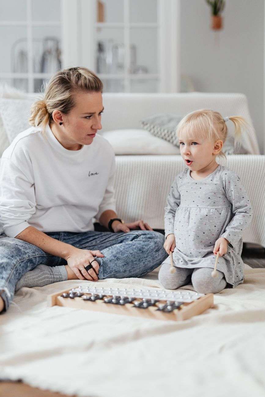 An adult sits on the floor with a young child as she plays a wooden xylophone with two mallets