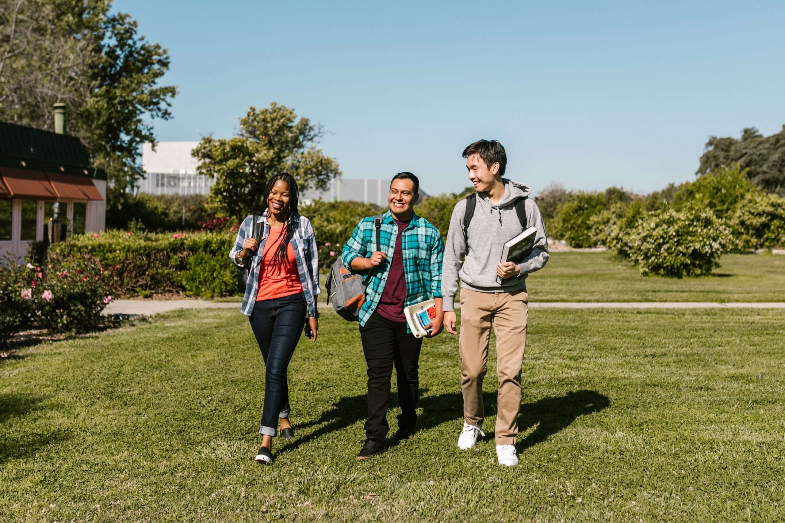 Three college students with backpacks and a notebook walking together across a grassy campus lawn on a sunny day