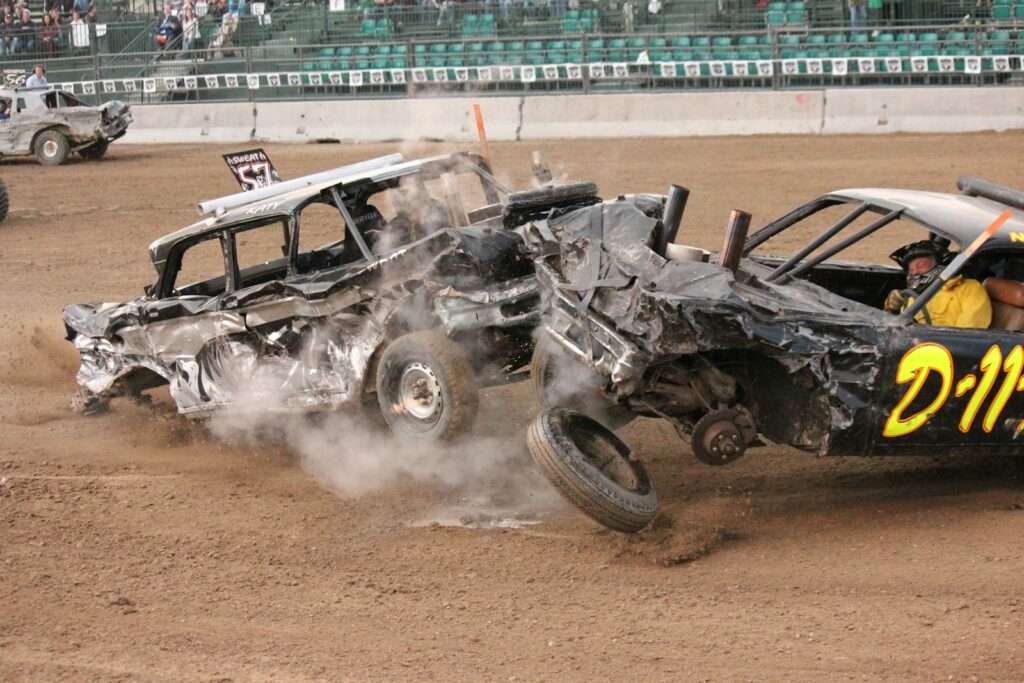 Two damaged cars colliding in a demolition derby arena, kicking up dust in front of a grandstand