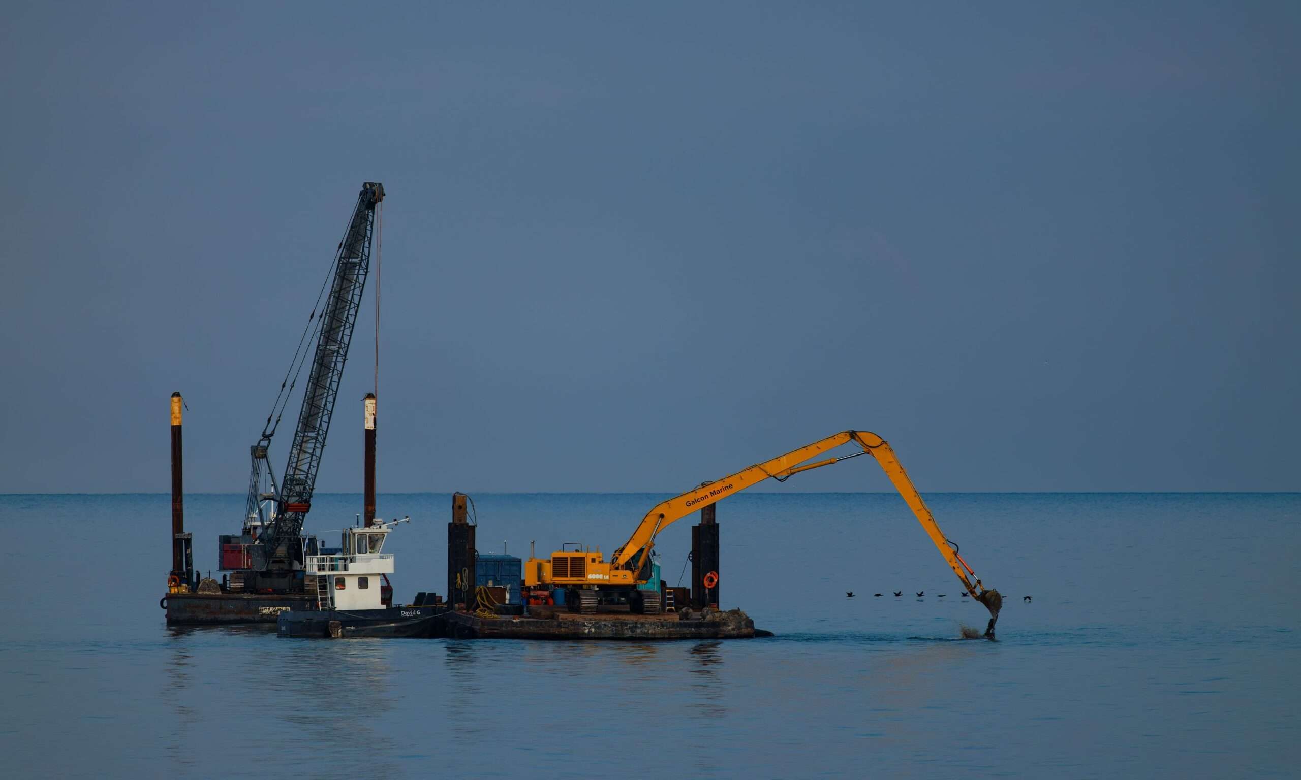 A crane barge with a long-reach excavator dredging sediment from open water under a clear sky