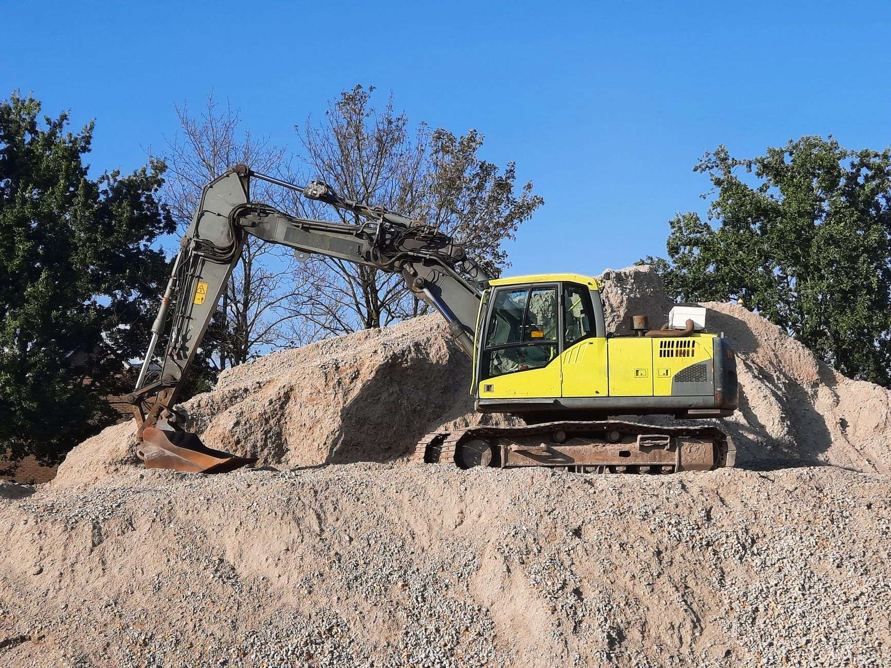 A yellow excavator parked on a large pile of sand and gravel under a blue sky