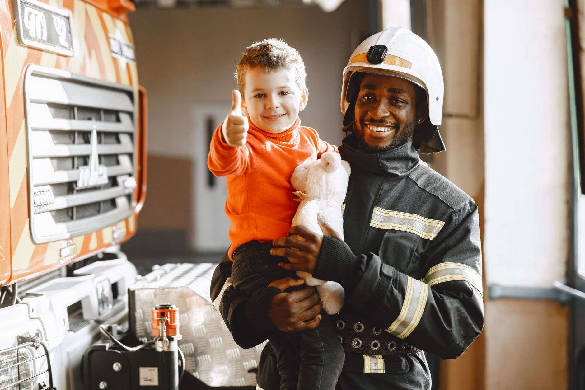 A firefighter in uniform and helmet smiles while holding a young child next to a fire engine at a fire station.