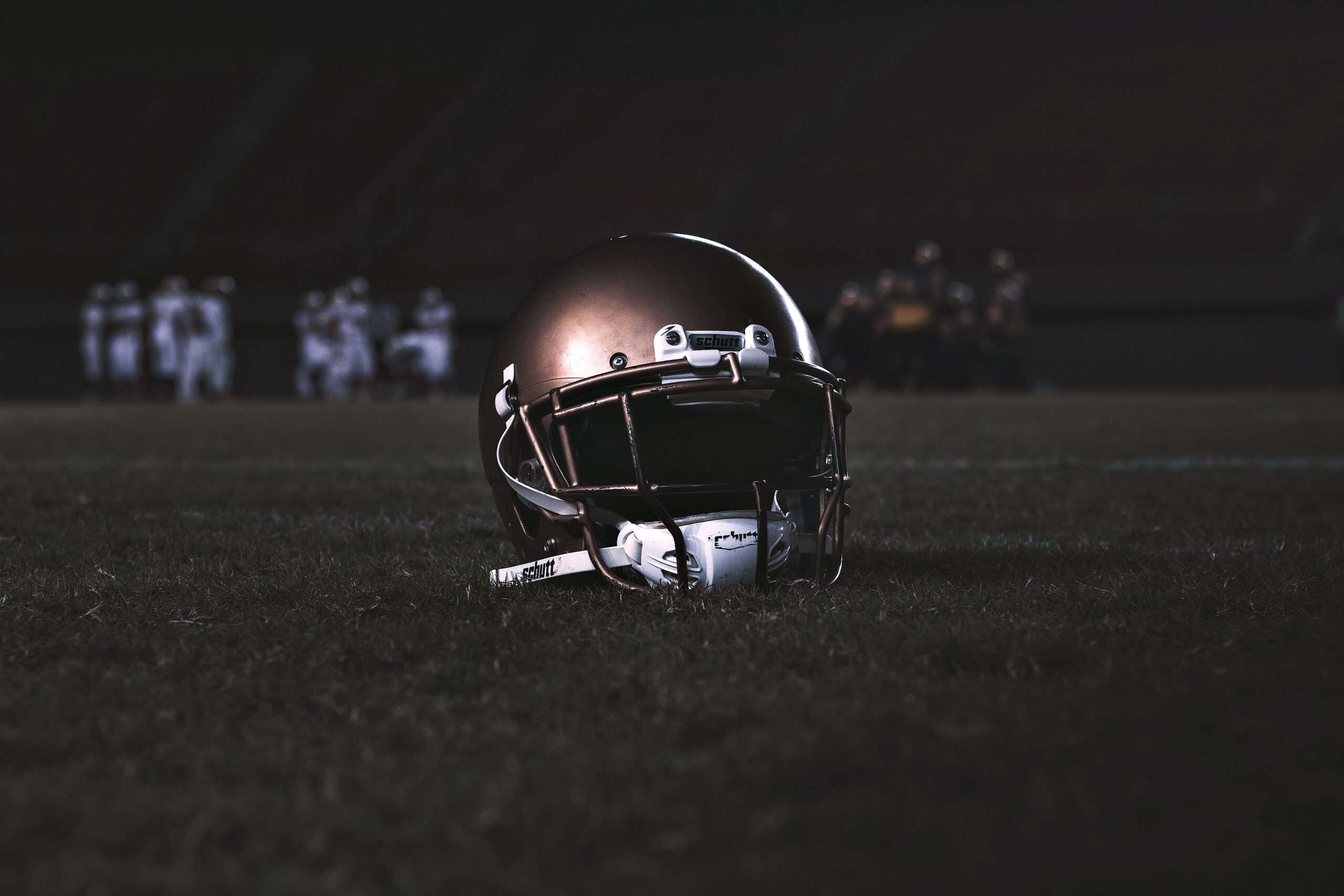 Football helmet resting on a grass field at night with players blurred in the background