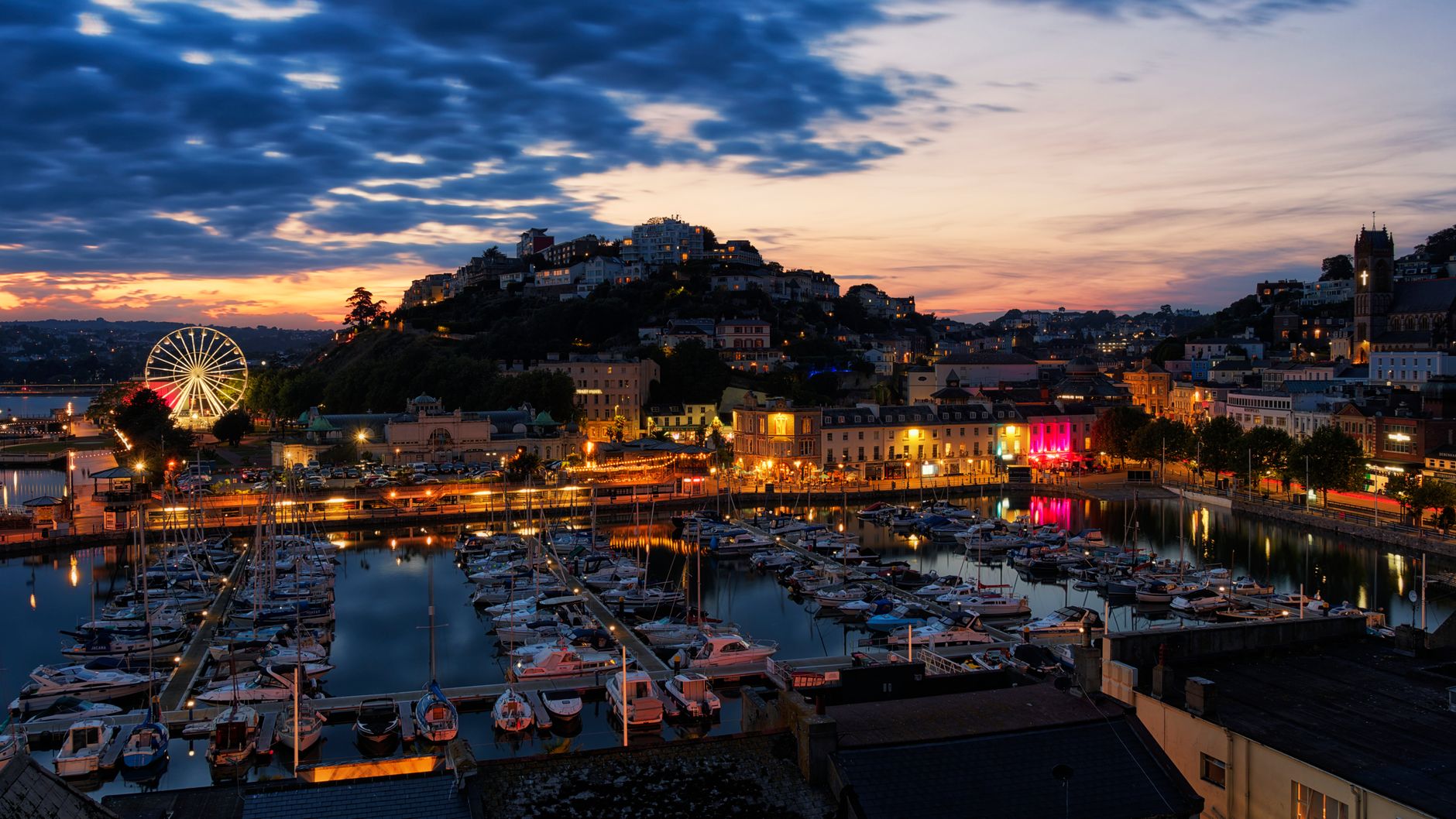 Boats docked at a waterfront marina with a lit Ferris wheel in the background at dusk