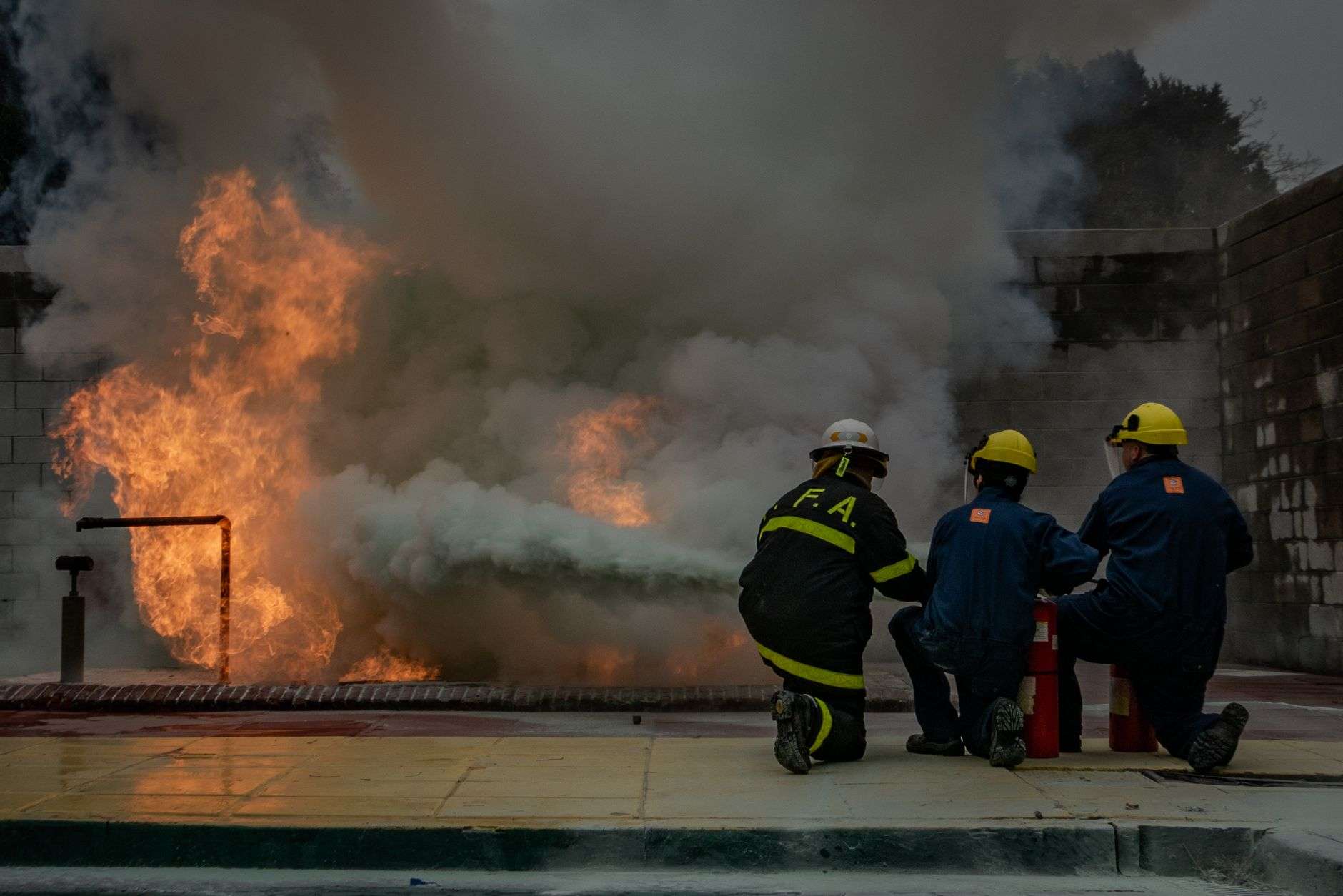 Firefighters in protective gear kneel near a controlled live-fire training blaze