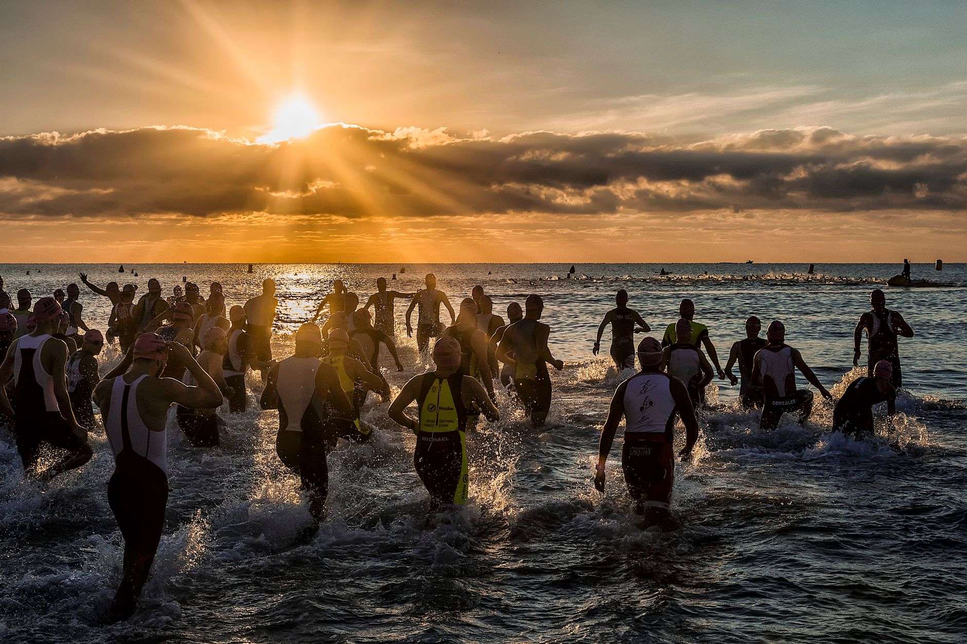 A group of triathletes in wetsuits and swim caps running into open water at the start of a race