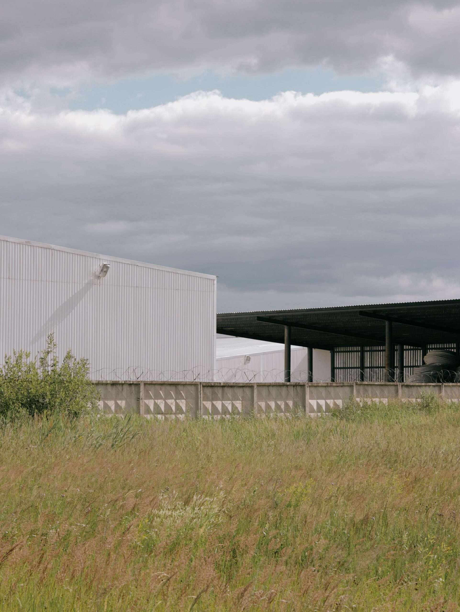 Plain industrial warehouse building behind a fence in an open grassy field under a cloudy sky