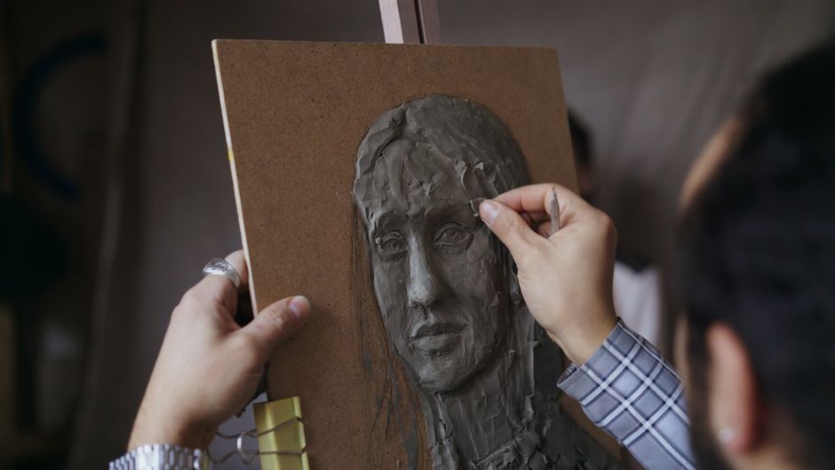 An artist's hands shaping a clay figure at a sculpture studio workbench
