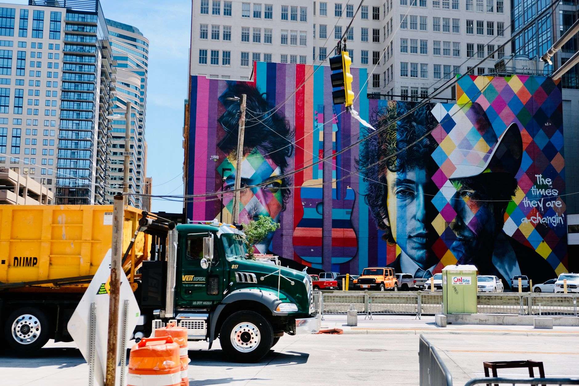 An artist on a lift painting a large colorful mural on the side of a downtown building