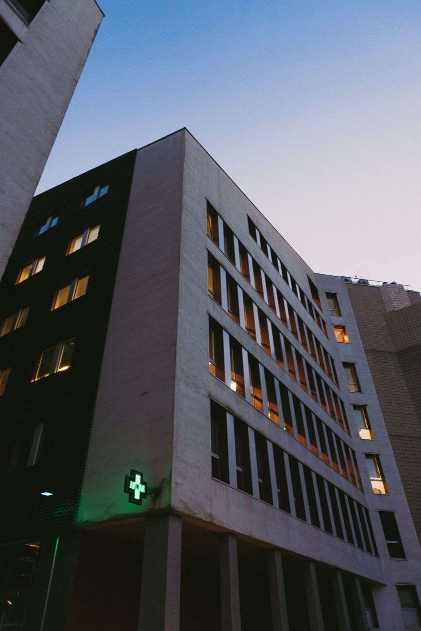 Low-angle view of a modern hospital building exterior at dusk with a lit green medical cross sign