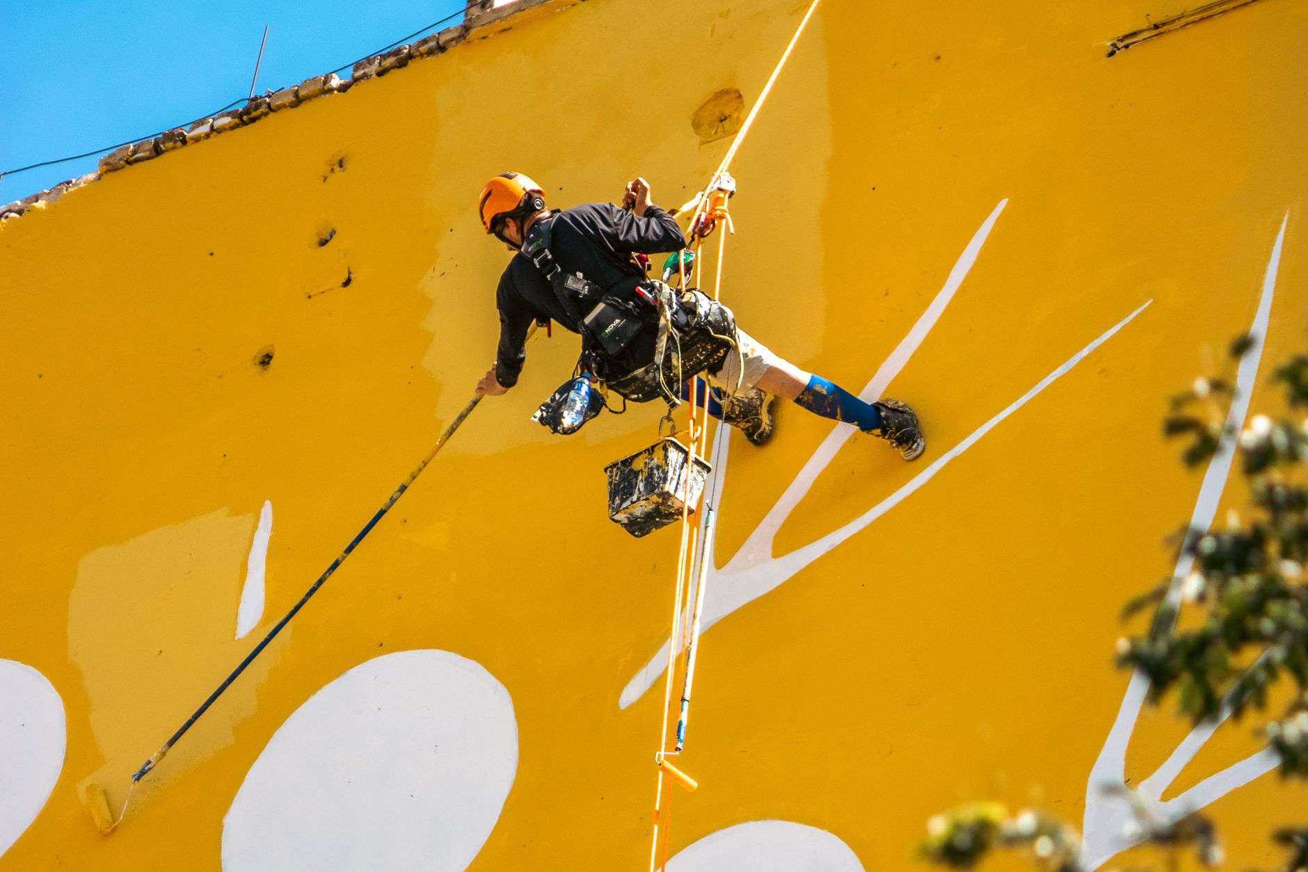 An artist standing on scaffolding painting a large colorful mural on an exterior wall
