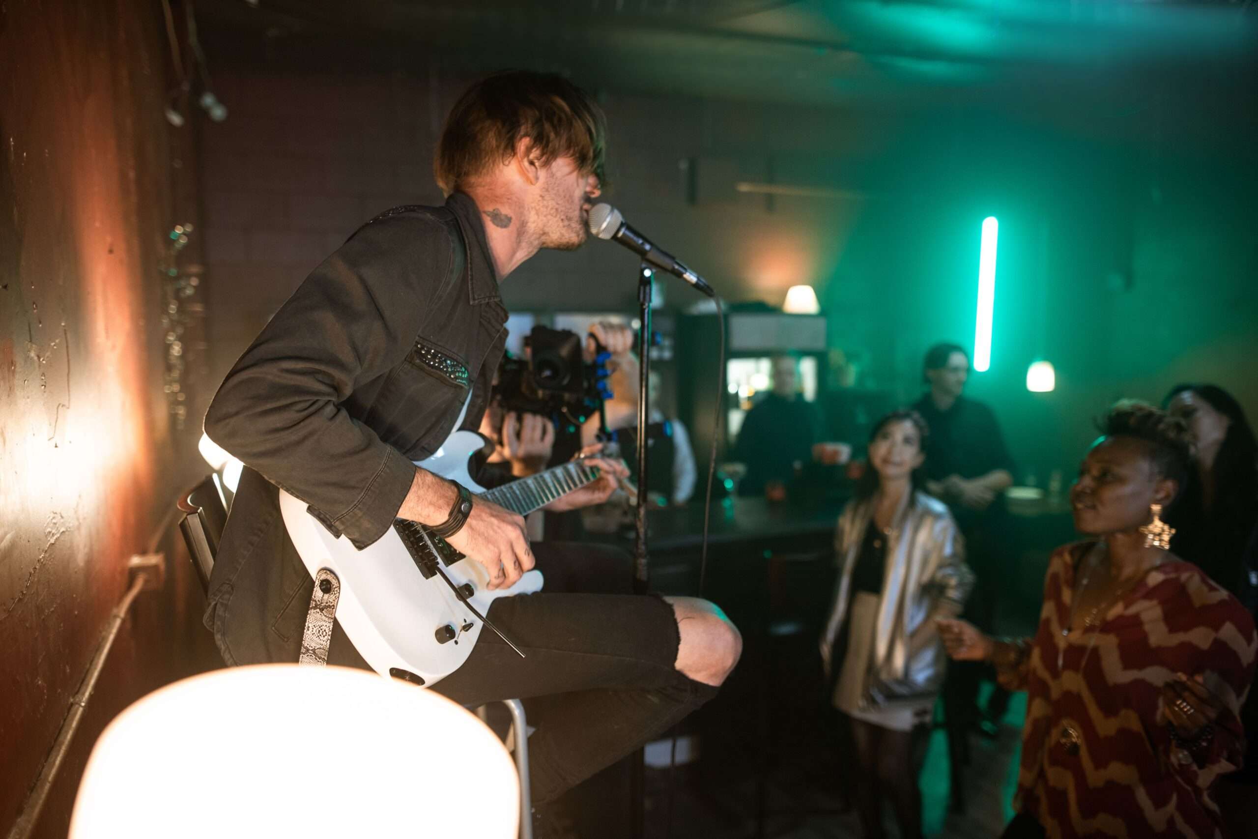 Musician playing an electric guitar and singing into a microphone at a small, intimate bar venue with onlookers nearby