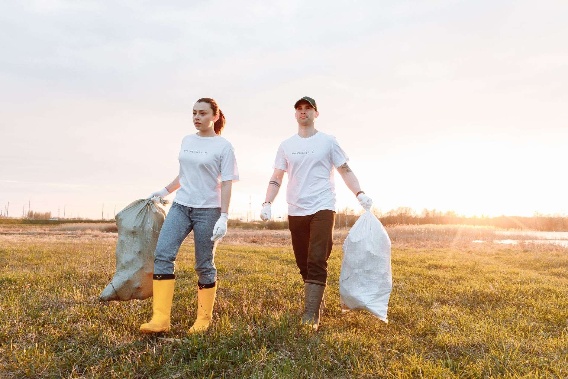 Two volunteers carrying bags of collected litter across a grassy field at sunset