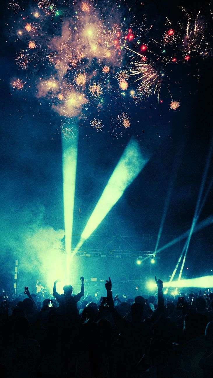 Crowd watching fireworks and stage lights at a nighttime outdoor celebration