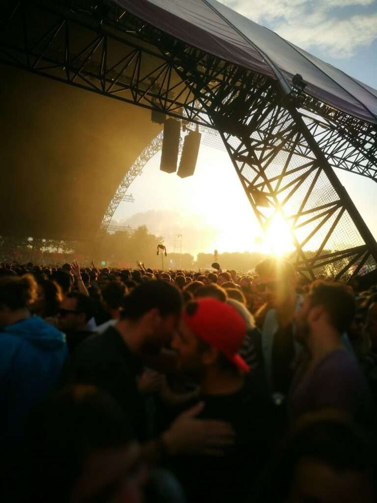 Crowd of concertgoers watching a performance at an outdoor music festival at sunset, with a stage and speaker rigging visible.