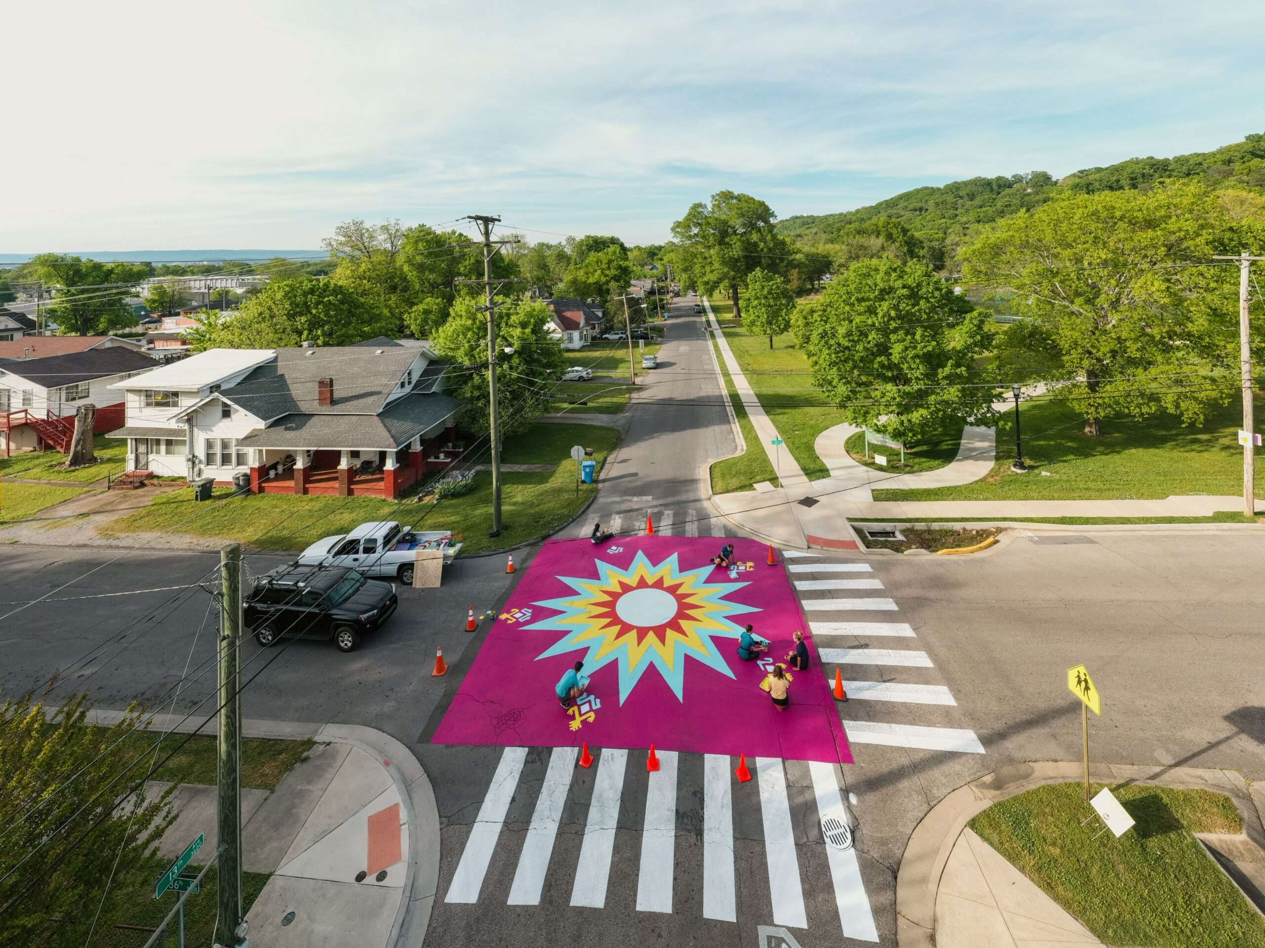 Aerial view of a colorful sunburst mural painted across a street crosswalk, with volunteers finishing the artwork near orange safety cones