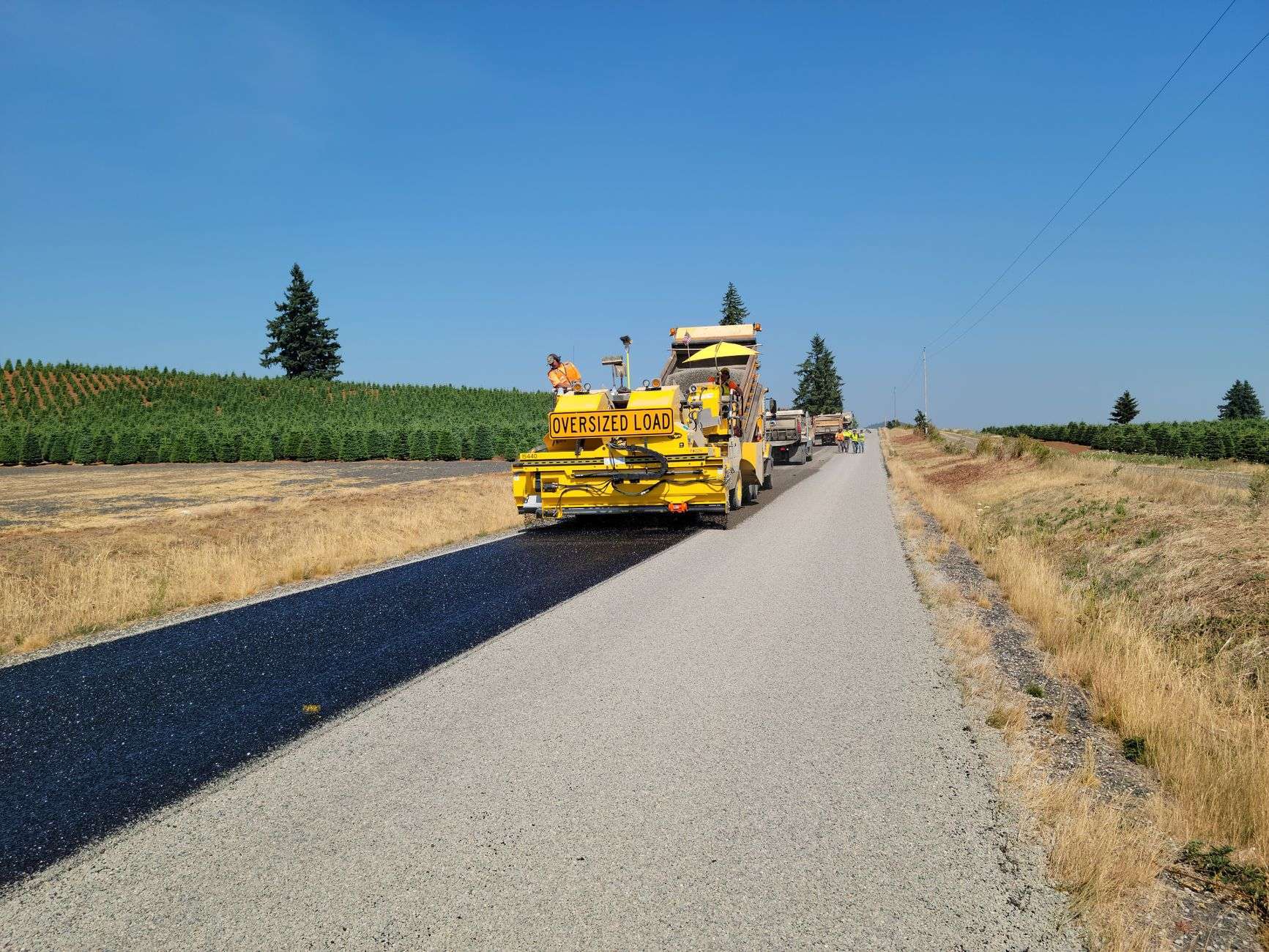 Road crew operating a paving machine to resurface a rural two-lane road with farmland on either side