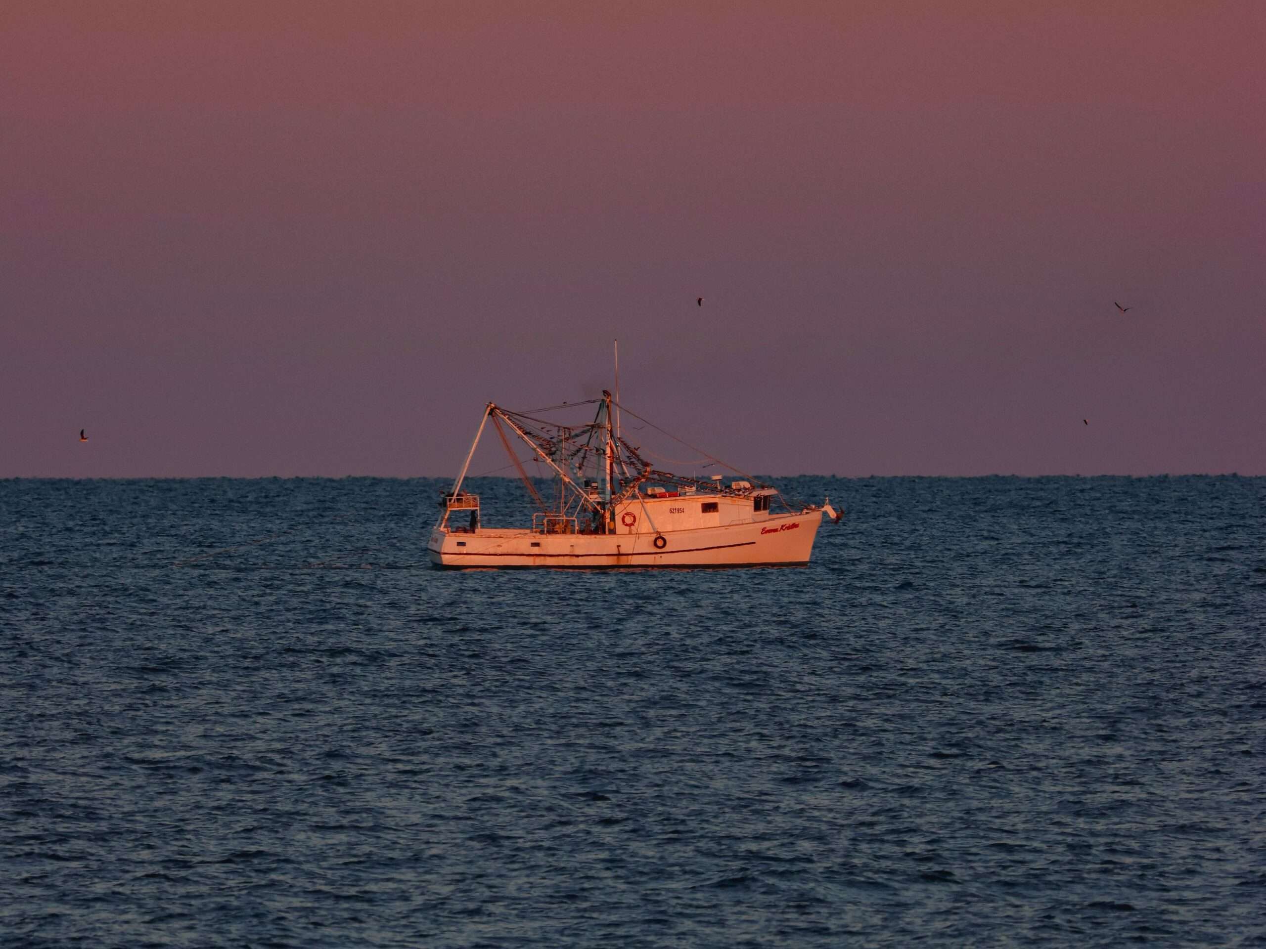 A shrimp trawler at sea silhouetted against a colorful sunset sky