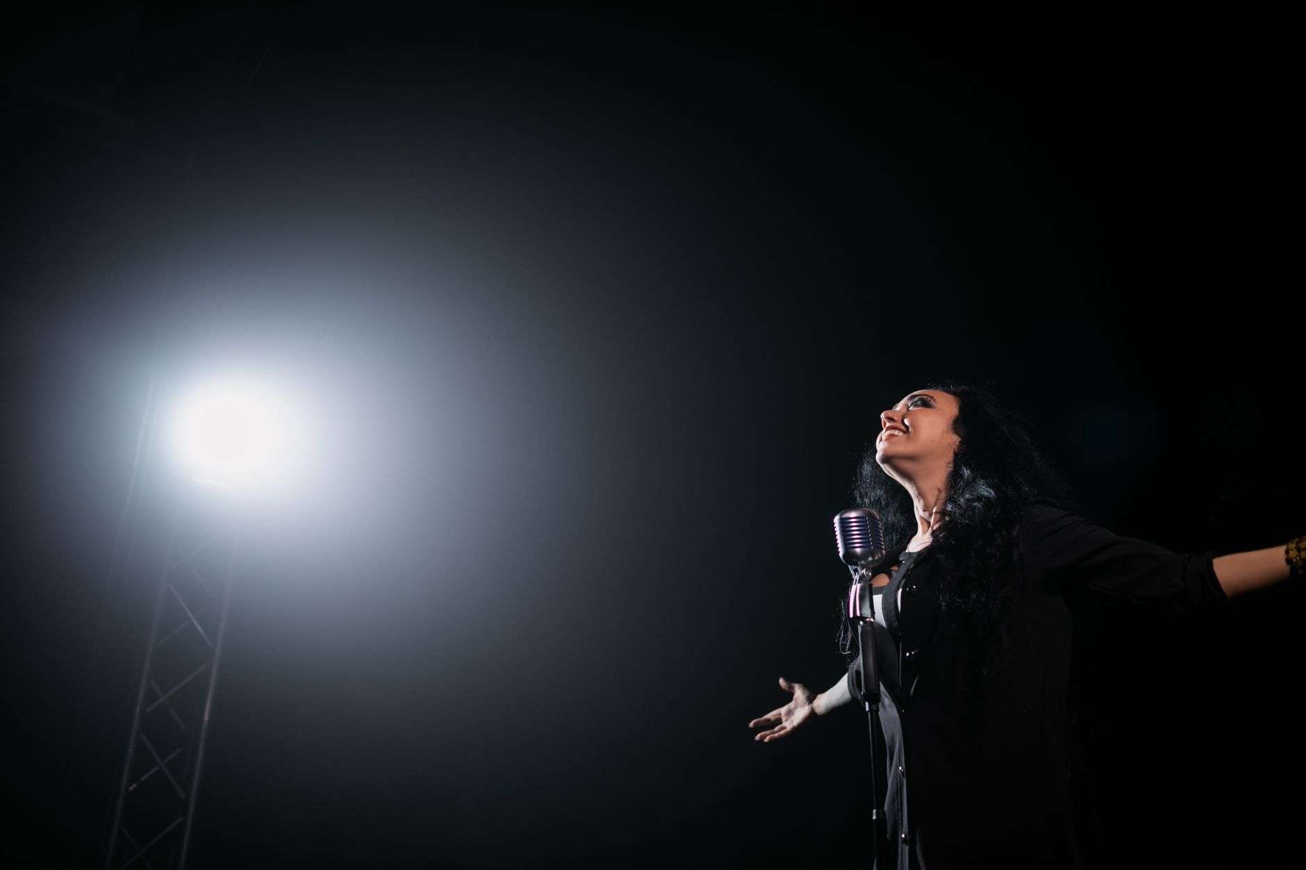 A woman with dark curly hair singing at a vintage microphone on a dark stage with a bright spotlight