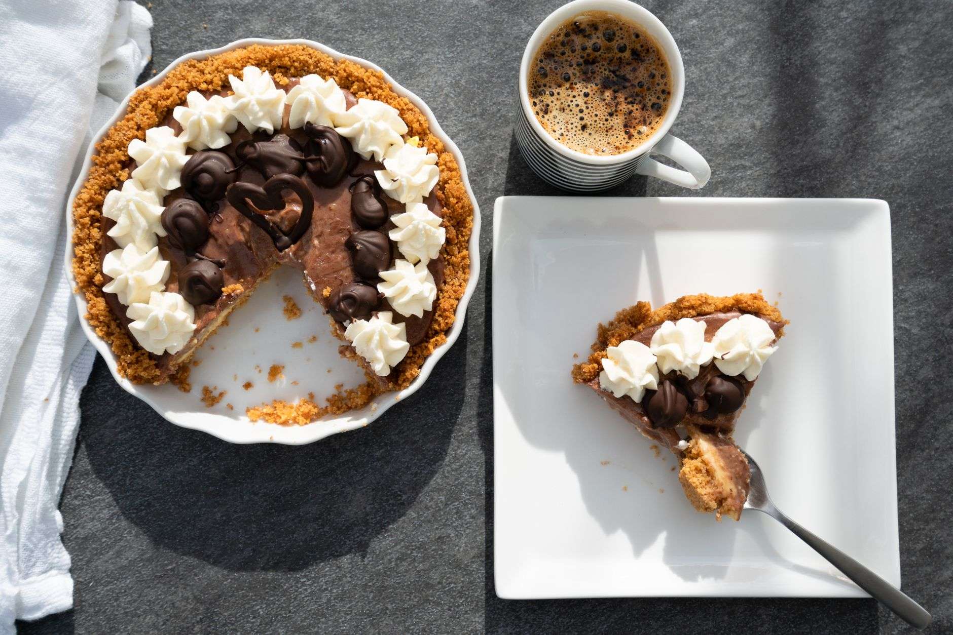 A chocolate pie with a cookie crust and whipped cream, with a slice on a plate and a cup of coffee