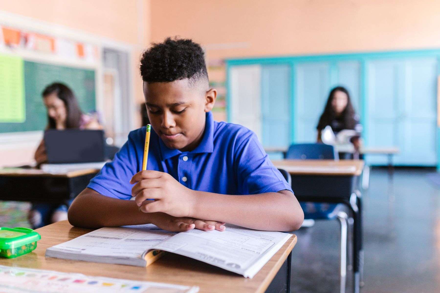 Student writing in a notebook at a classroom desk while other students work in the background