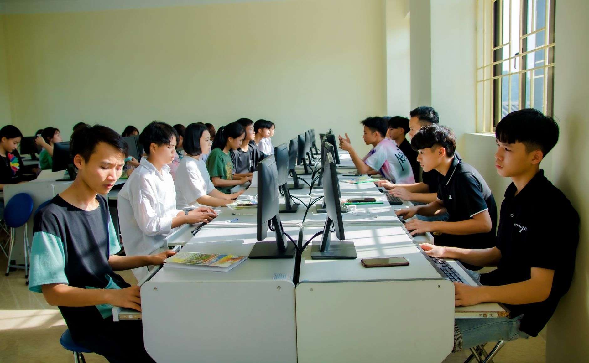 Students seated at rows of desktop computers in a sunlit classroom