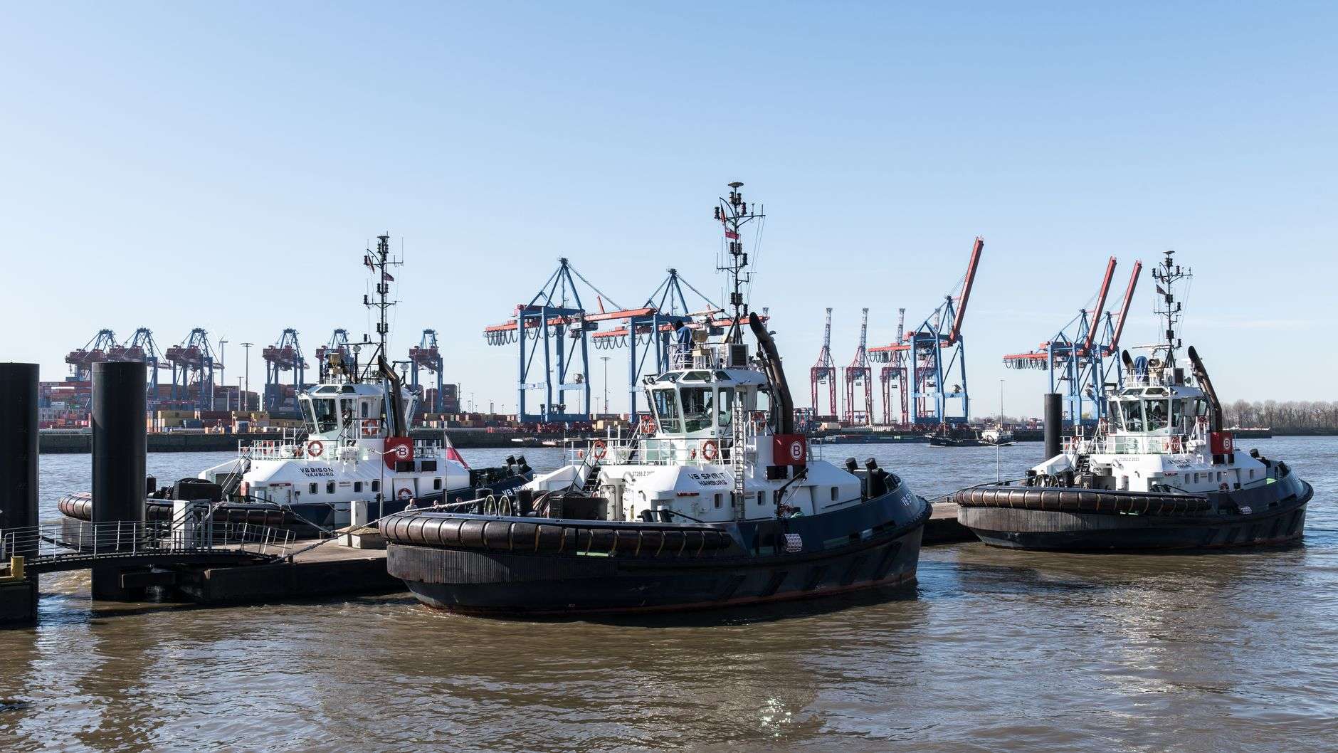 Three tugboats moored in a harbor with container cranes in the background