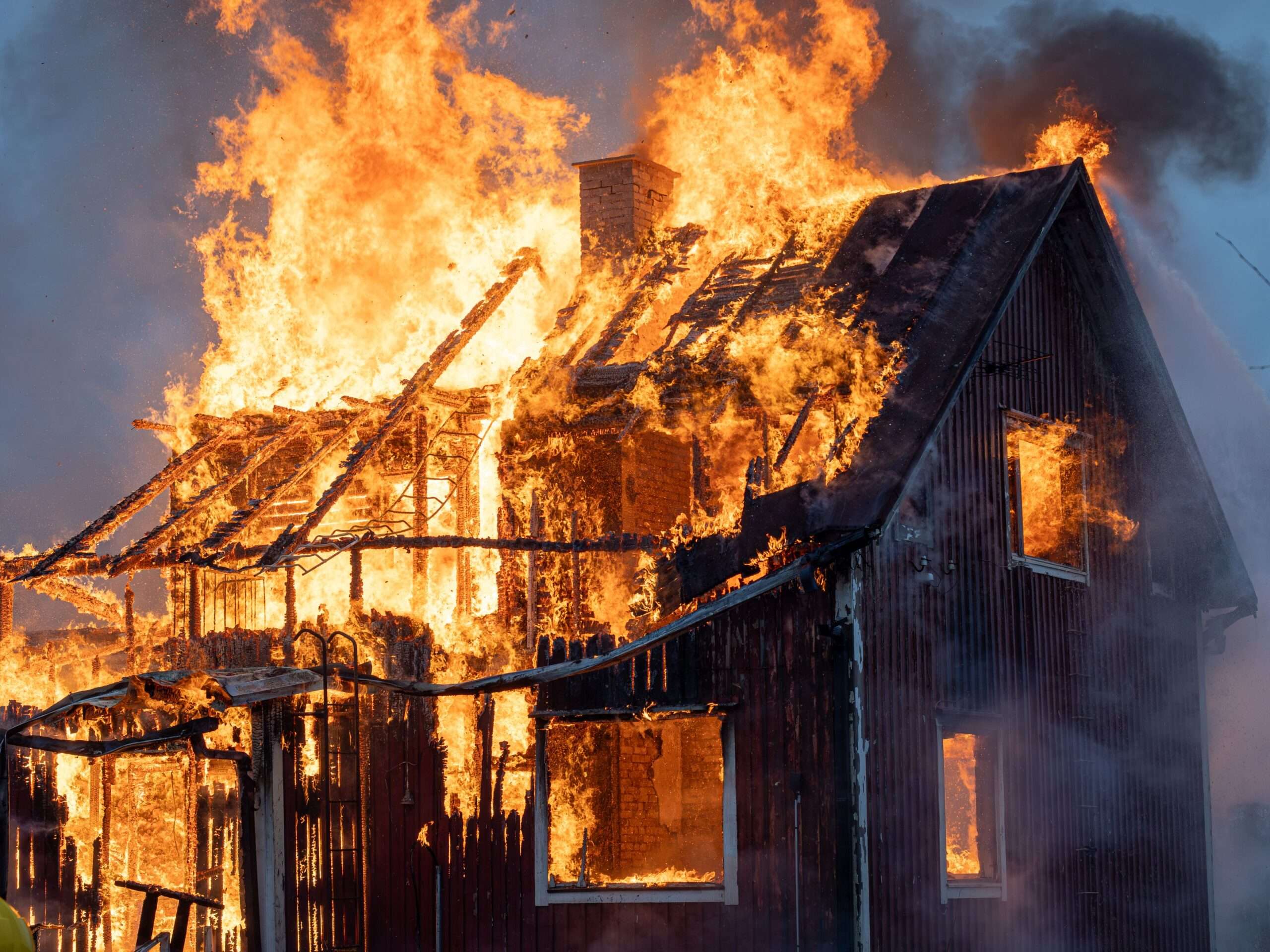 A wood-frame building fully engulfed in flames at night, with heavy fire and smoke coming from the roof and windows