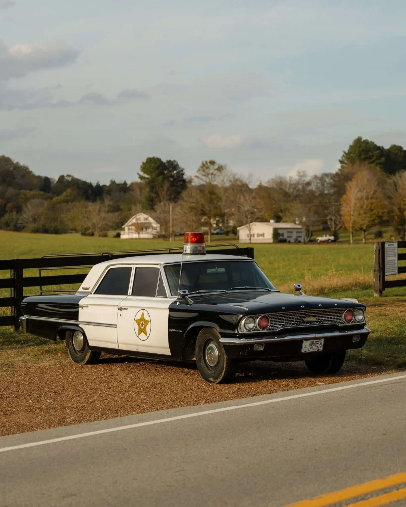 Vintage black-and-white police cruiser with a rooftop light parked along a rural roadside, with farmland in the background