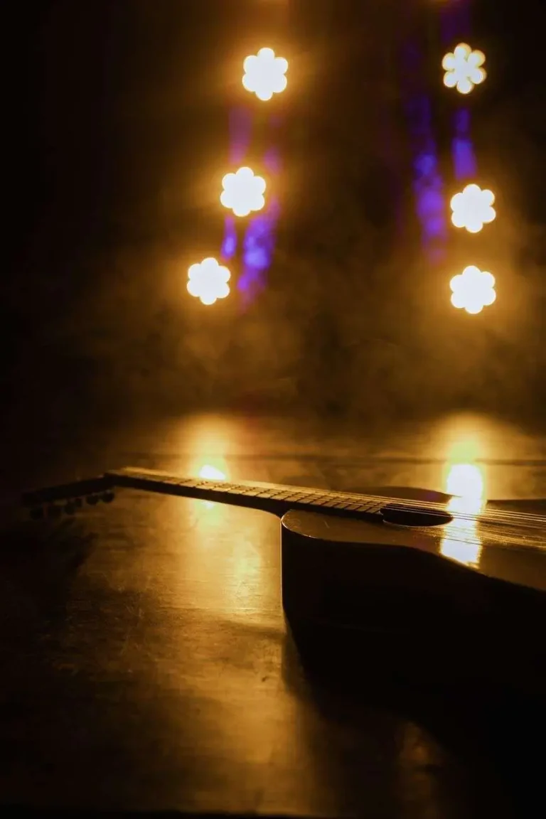 A single acoustic guitar resting on a stage floor, lit by warm spotlights against a dark, hazy backdrop
