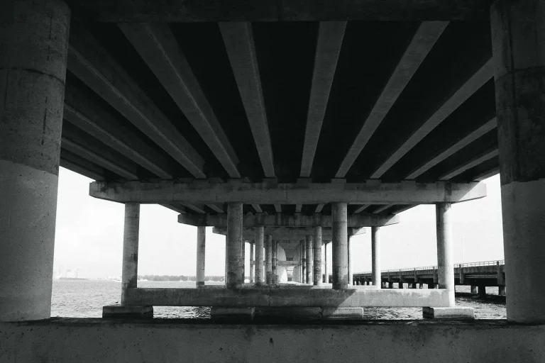 View from the water looking up at the underside of a concrete highway bridge, showing its support pilings and roadway deck