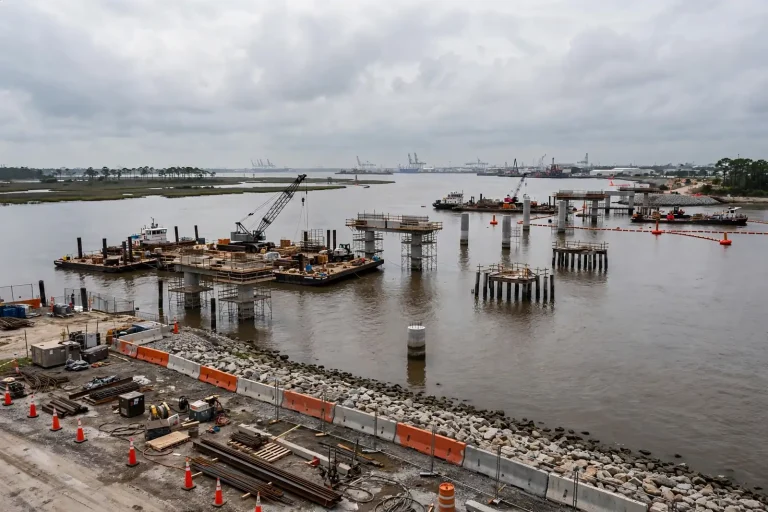 Interstate highway bridge over a river with cargo cranes and a city skyline in the background