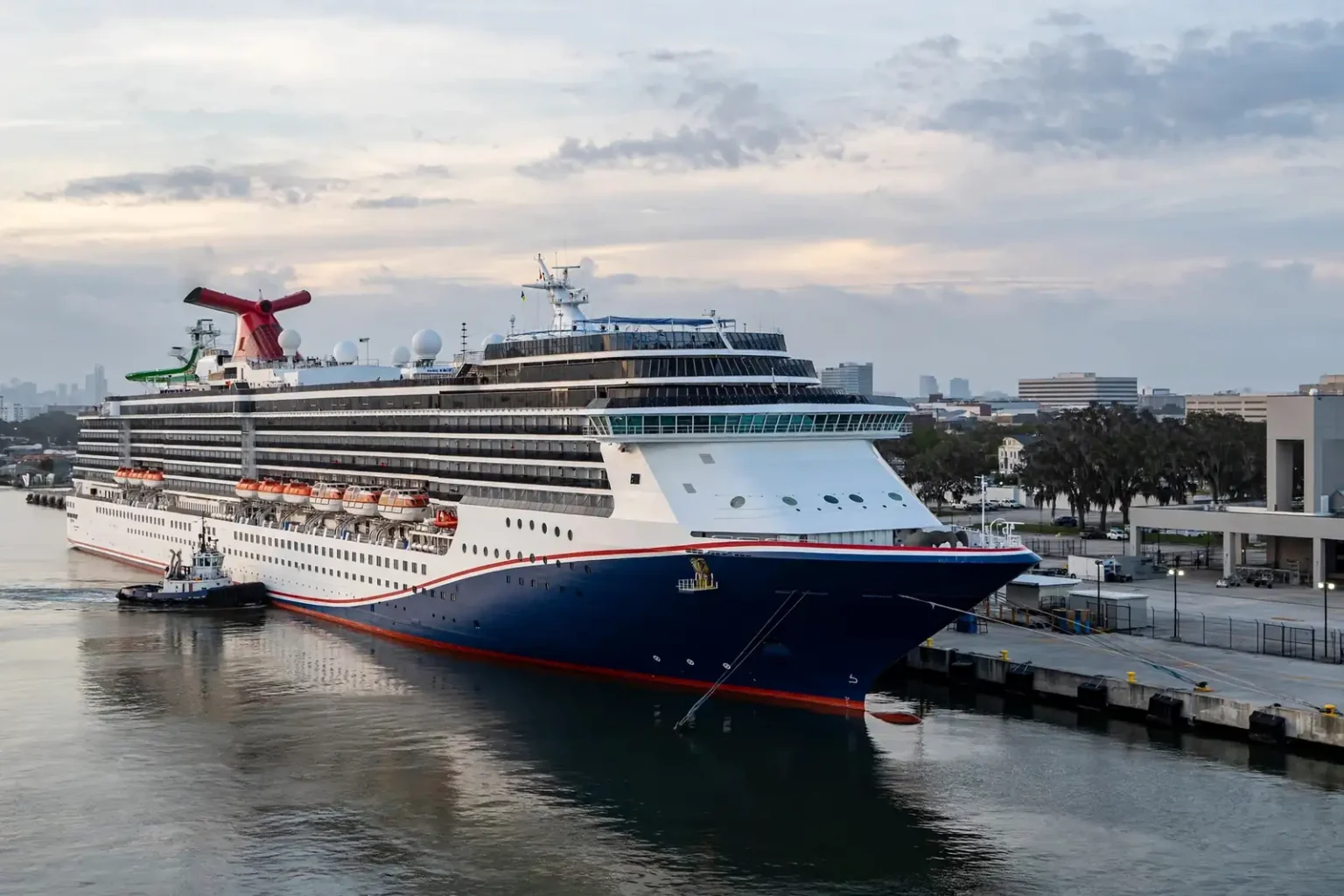 A white Carnival cruise ship with a red-and-blue funnel docked beside the cruise terminal on the Mobile River