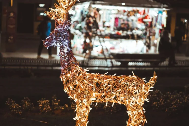 A lighted wireframe reindeer decoration glowing at night with an illuminated holiday market stall in the background