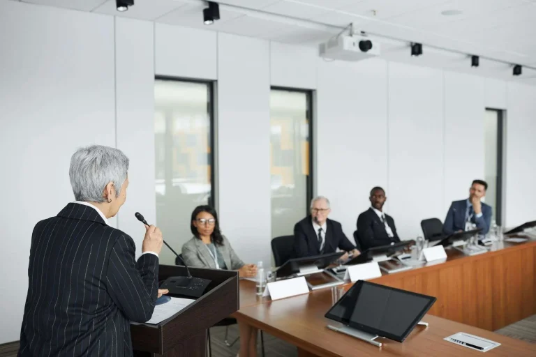 Speaker at a podium with a microphone addressing a panel of seated officials in a meeting room