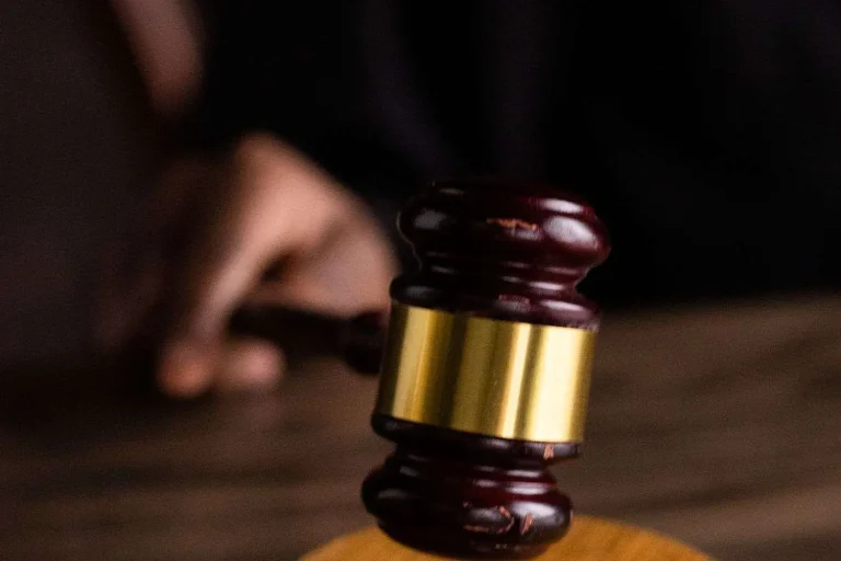 A wooden judges gavel with a brass band, resting upright on a sound block in a dim courtroom, with a blurred hand in the background