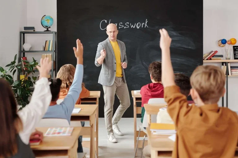 A teacher standing at a chalkboard leads a classroom of students, several with hands raised