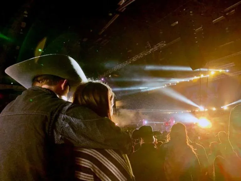 Concertgoers wearing cowboy hats watch a brightly lit stage at a nighttime outdoor country music concert
