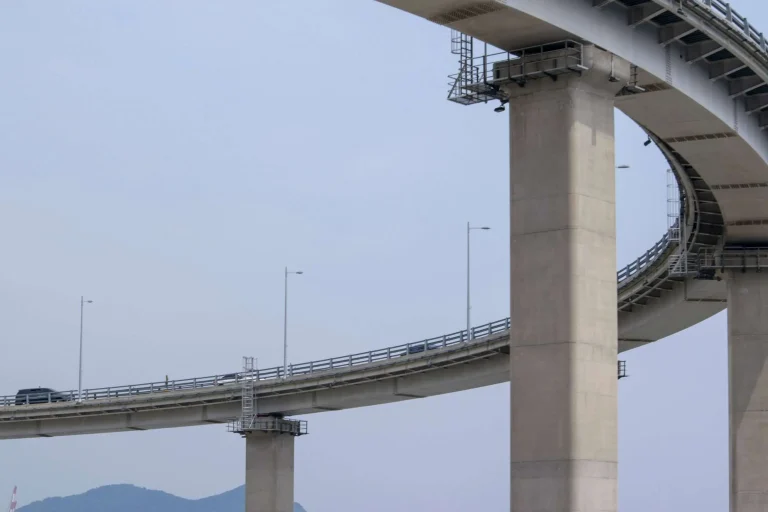An elevated concrete highway bridge on tall support piers curves above open water and low hills.