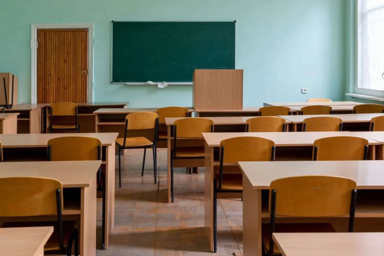 Rows of empty wooden desks and chairs in a classroom with a chalkboard on the wall.
