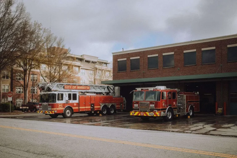 Two red fire trucks parked side by side in front of a brick fire station on a cloudy day