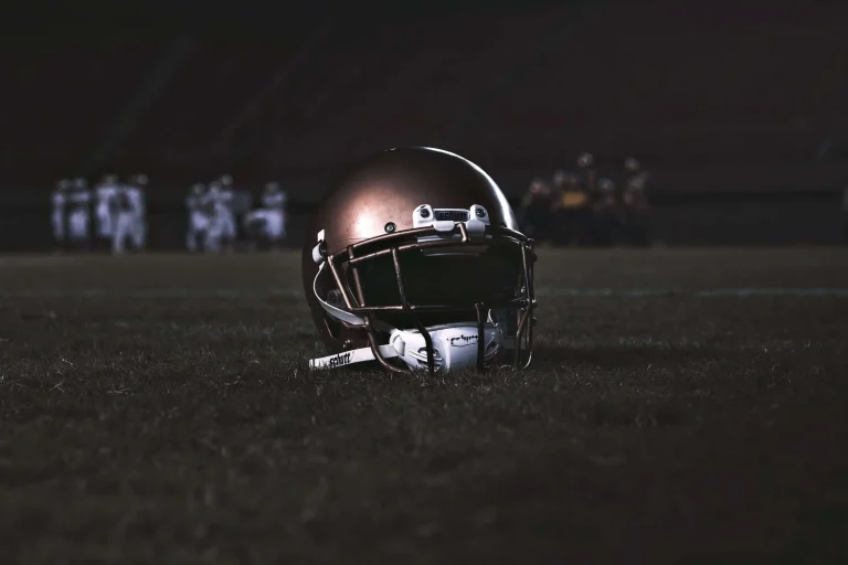 Football helmet resting on a grass field at night, with players blurred in the background