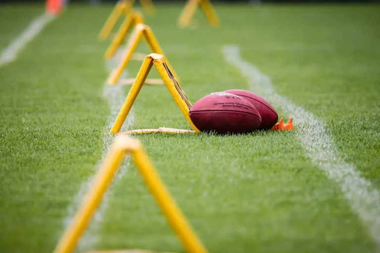 An American football resting on a grass practice field next to yellow training hurdles