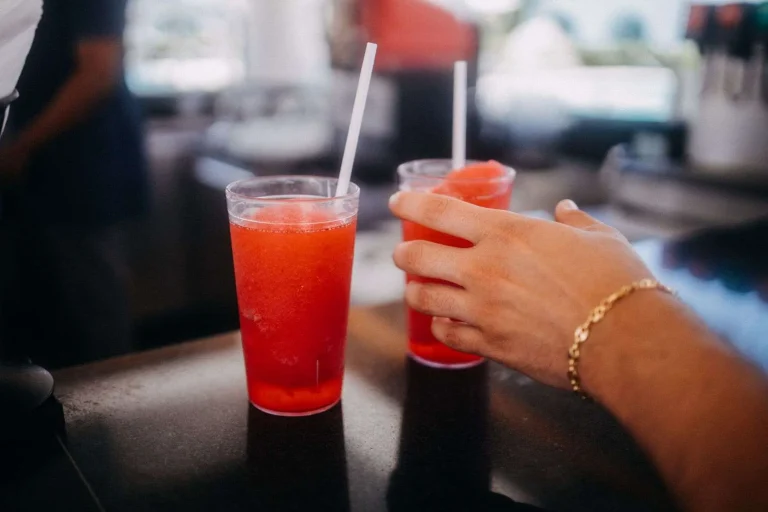 Two plastic cups of red frozen drinks with straws on a bar counter, with a hand reaching for one