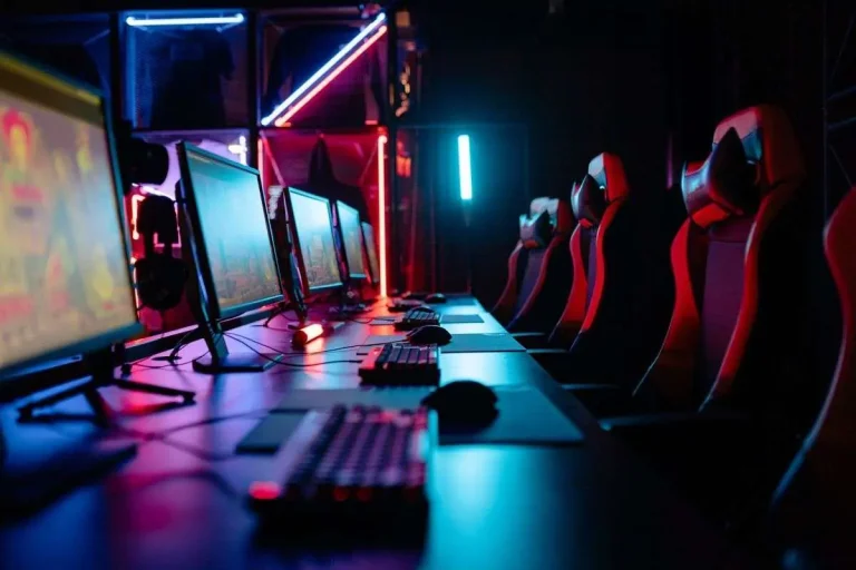 Row of gaming computers with illuminated monitors and red gaming chairs under neon lighting