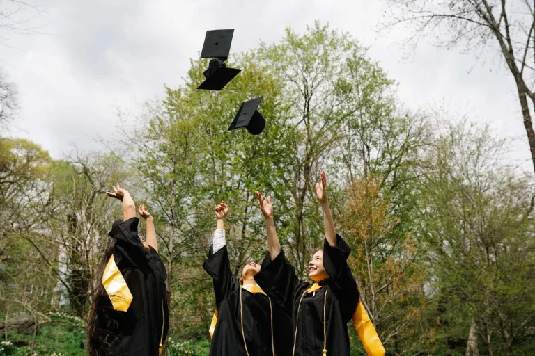 Three graduates in black caps and gowns tossing their graduation caps into the air outdoors