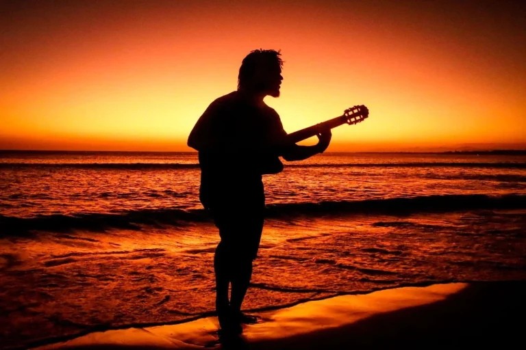 Silhouette of a barefoot man playing a guitar at the edge of the water during a beach sunset